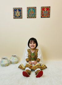 Child in traditional outfit sitting between two decorative vases with wall art in the background.