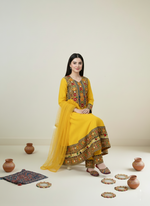 Woman in a yellow traditional outfit with intricate patterns, standing against a plain background.