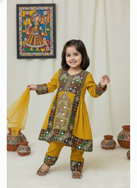 Child wearing a yellow traditional outfit with intricate patterns, standing against a white curtain with decorative pots and a wall hanging.