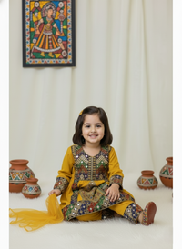 Young girl in a traditional outfit sitting on a white floor with decorative pots and a colorful wall hanging.