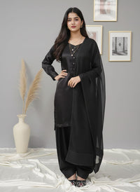 A woman modeling a black three-piece traditional outfit with a matching sheer dupatta, standing against a gray wall with framed art and a decorative vase.