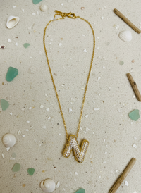 Gold necklace with letter 'N' pendant on a textured surface with shells and driftwood.