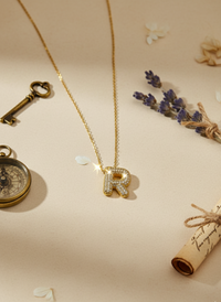 Gold necklace with 'R' pendant on a decorative background with a key, compass, and rolled paper.