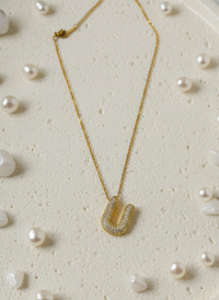 Gold necklace with a U-shaped pendant on a textured surface with scattered pearls