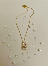Gold necklace with a letter 'S' pendant on a beige background with scattered pearls and crystals.