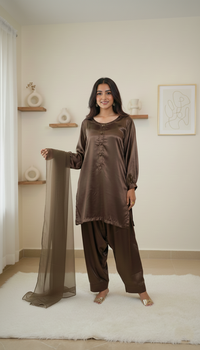 A woman modeling a brown satin kurta and pant set with a matching sheer dupatta in a bright, modern room with beige walls and white decor.