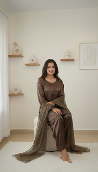 A woman wearing a brown satin kurta set with a matching dupatta sits on a white stool in a minimalist, warm-toned room with wooden floating shelves and a white rug.