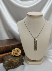 Necklace with a decorative pendant on a mannequin bust, next to a rose and ornate box on a white fabric background.