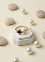 Gold rings with black and gold accents on a white box surrounded by pearls on a beige background