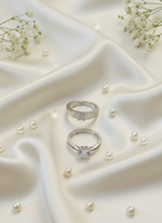 Two silver rings with gemstones on a white fabric background with scattered pearls.