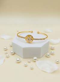 Gold bracelet with decorative element on a white hexagonal base surrounded by pearls on a beige background