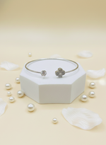 Silver bracelet with clover charm on a white hexagonal stand with pearls and petals on a beige background