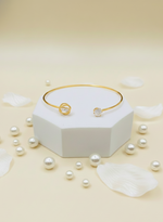 Gold bracelet with pearls on a white hexagonal stand and scattered pearls on a beige background