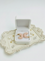 Pink crescent moon earrings in a white jewelry box on a decorative tray.