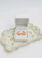 Orange earrings in a white jewelry box on a decorative tray with pearls.