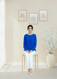 Woman in a blue top and white pants sitting on a chair in a room with light-colored walls and decor.