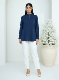 Woman wearing a navy blue top with white pants standing in a minimalistic setting with a decorative plant.