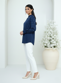 Woman wearing a navy blue top with white pants standing next to a vase of flowers.