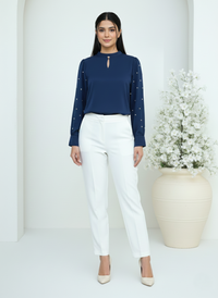 Woman wearing a navy blue top with white polka dots and white pants, standing against a white background with a decorative plant.