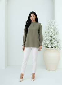 Woman wearing a green top and white pants standing against a white background with a decorative vase and flowers.