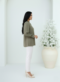 Woman in a green sweater and white pants standing next to a large vase with flowers in a minimalistic setting.