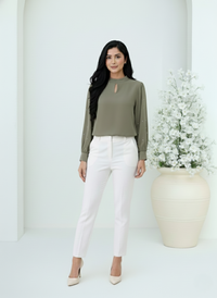 Woman wearing a green top and white pants standing in a minimalistic setting with a vase of flowers.