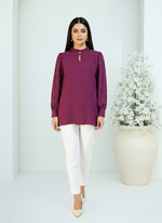 Woman wearing a purple top and white pants standing against a white background with a decorative vase.