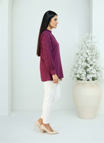 Woman wearing a purple blouse and white pants standing against a white background with a decorative plant.