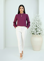 Woman wearing a purple blouse and white pants standing next to a vase with flowers on a white background