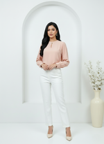 Woman wearing a pink blouse and white pants standing in a minimalistic setting with a vase and flowers.