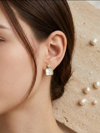 Close-up of a person wearing gold square earrings with pearls on a beige background