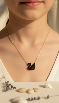 Gold necklace with a black swan pendant worn by a person, with seashells and lavender in the background.