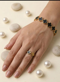 Hand wearing a gold bracelet and ring with black clover designs, surrounded by pearls on a beige background.