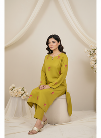 Woman in a mustard yellow outfit with red patterns sitting on a white stool against a beige curtain background.