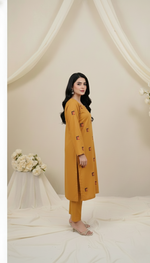 Woman wearing a mustard yellow outfit with a long coat and pants in a softly lit room.