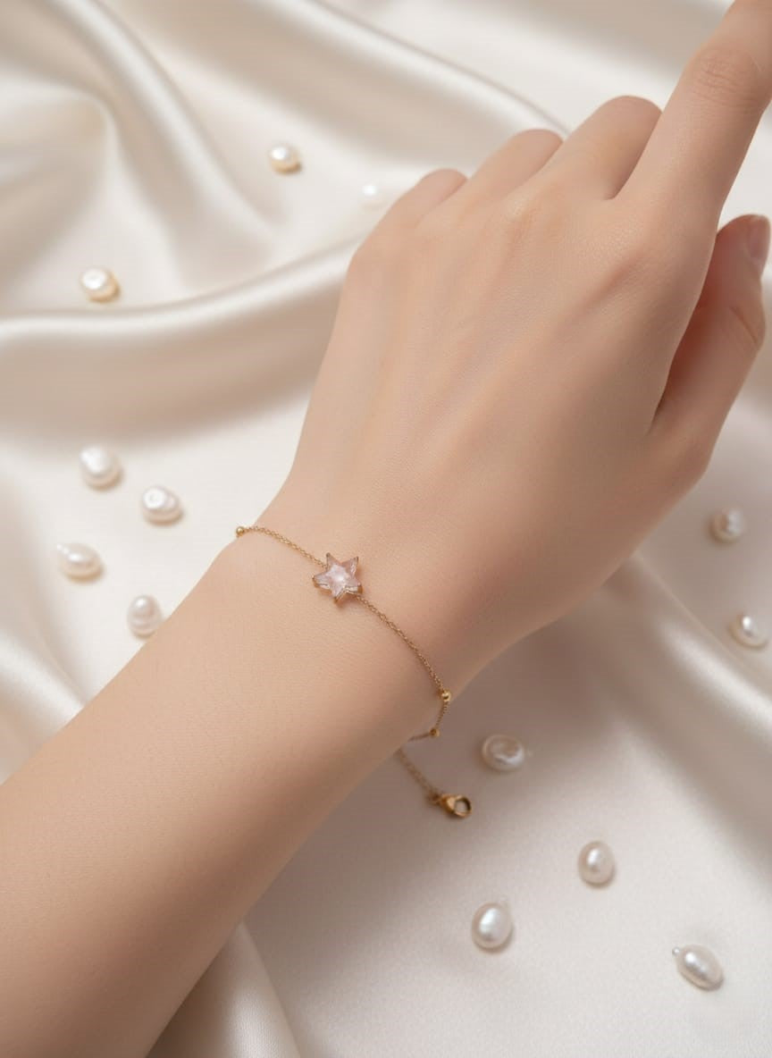 Hand wearing a gold bracelet with a pink flower charm on a white fabric background.