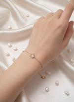 Hand wearing a gold bracelet with a pink flower charm on a white fabric background.