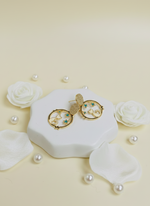 Gold earrings on a white hexagonal jewelry box with decorative flowers and pearls on a light yellow background