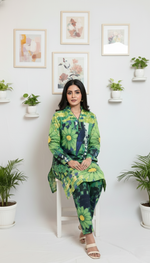 Woman in a green floral outfit sitting on a white stool against a white wall with framed pictures and plants.
