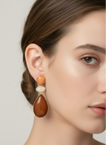 Close-up of a woman wearing a pair of earrings with orange and white stones on a neutral background