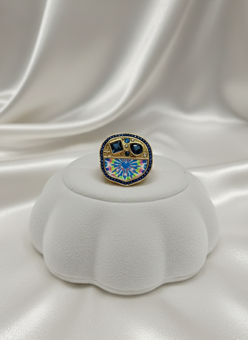 Decorative ring with blue and green gemstones on a white cushion against a white fabric background