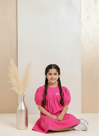 Young girl in a pink dress sitting on the floor with a vase and dried plants in the background.