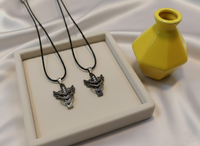 Two silver necklaces with pendants on black cords, next to a yellow vase on a white fabric background.