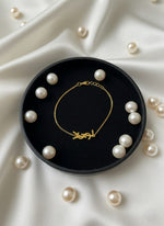 Gold bracelet on a black dish surrounded by pearls on a white fabric background
